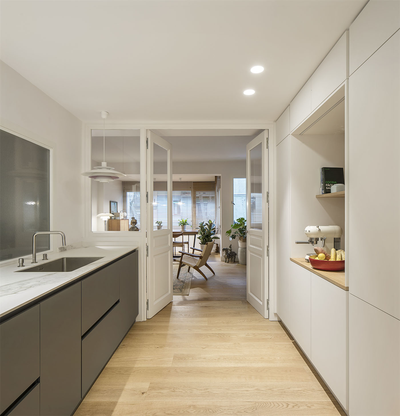 Imagen de una cocina con una encimera color blanca y muebles color gris debajo a un lado y en frente una pared armario con muebles altos color balnco que contienen la nevera, el horno, y demas electrodomesticos.